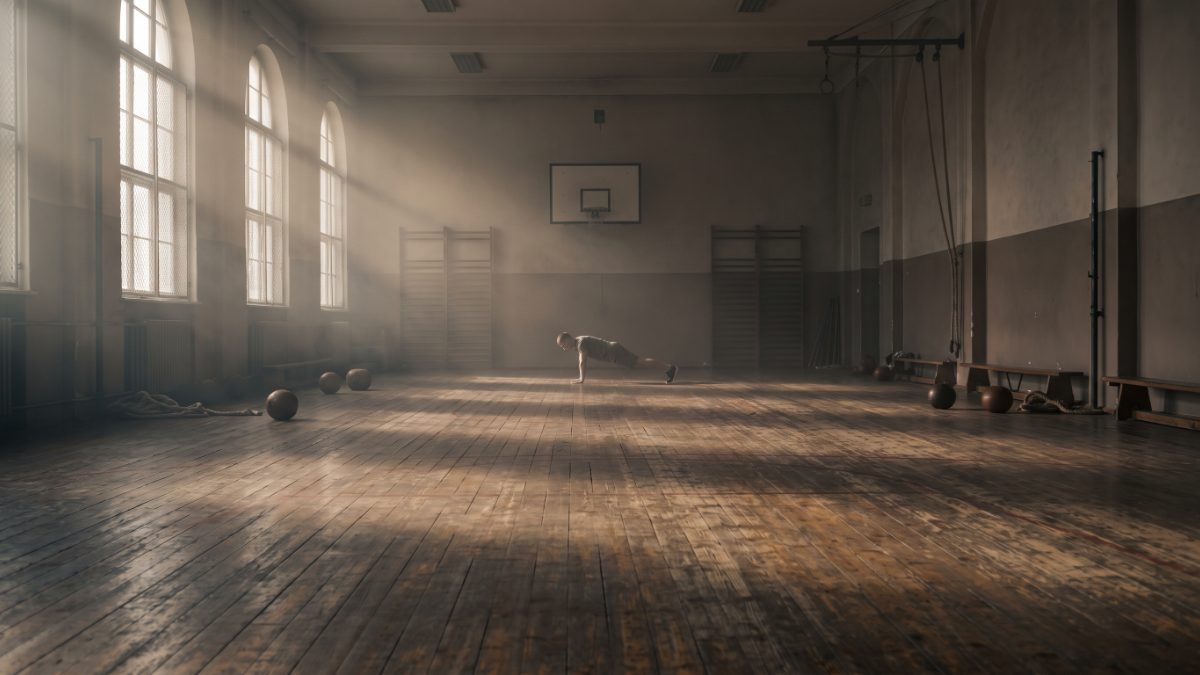 Alte Turnhalle mit hohen Bogenfenstern und abgenutztem Holzboden, weit hinten hält eine einzelne Person einen Liegestütz.